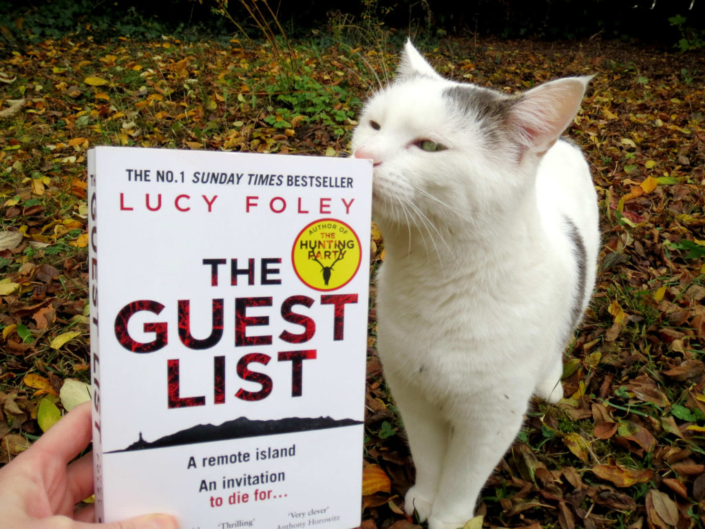 Im Vordergrund hält jemand "The Guest List" von Lucy Foley in die Kamera. Eine weiße Katze reibt ihre Nase am Buch. Im Hintergrund sieht man eine Wiese voller herblistlichem Laub.