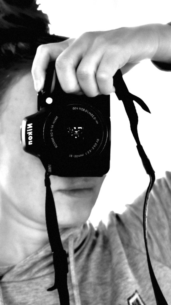 Ein Selfie von mir mit meiner Nikon im Spiegel, in Graustufen gehalten. Die Kamera verdeckt mein Gesicht.